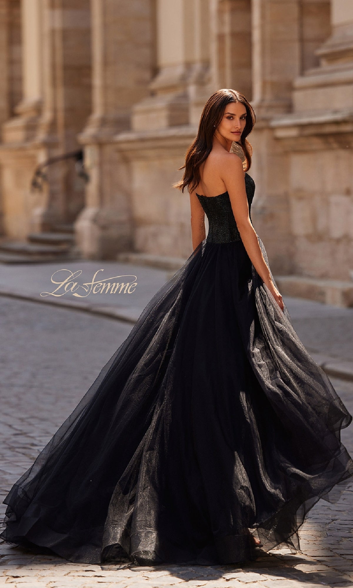 A woman in the La Femme 32690 Strapless Long Prom Ball Gown stands on a cobblestone street before a stone building, looking over her shoulder.